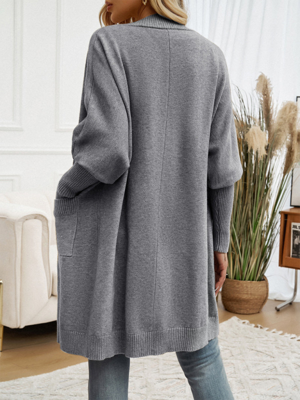 Woman wearing a gray batwing sleeve cardigan solid color sweater jacket, standing in a cozy room with minimalist decor.